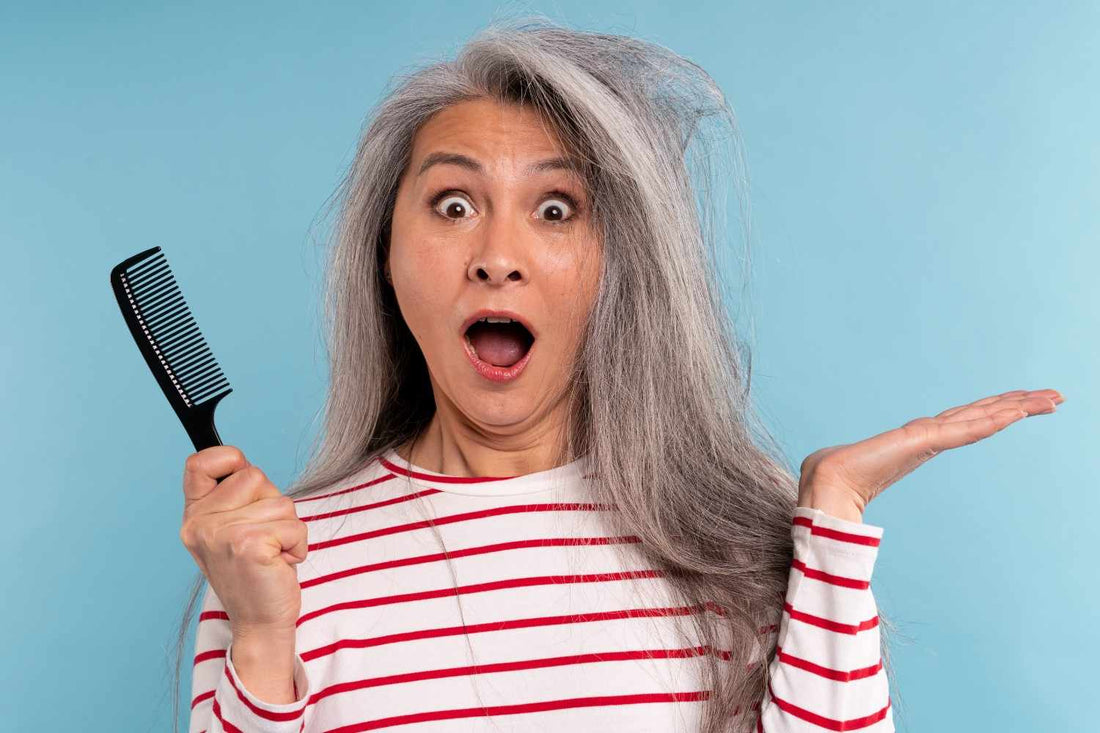 Surprised middle-aged woman holding a comb and looking at her tangled gray hair, symbolizing hair concerns related to hormonal changes. hair thinning treatment for women, gray hair care routine, hormonal imbalance hair loss